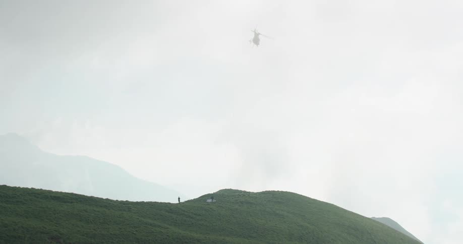 A helicopter flying in the sky with a hill underneath and mountains in the background