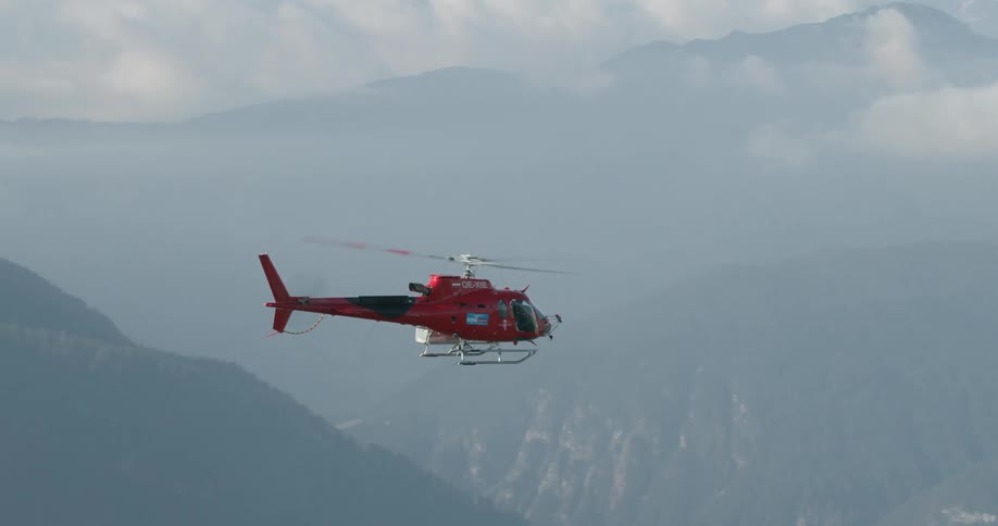 A red helicopter flying in the air with mountains and clouds in the background