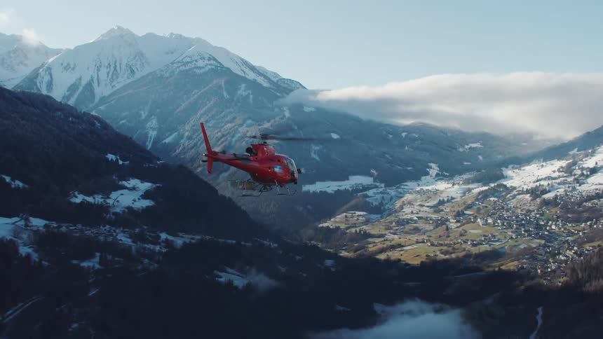 A red helicopter flies over snowy mountains in the shade