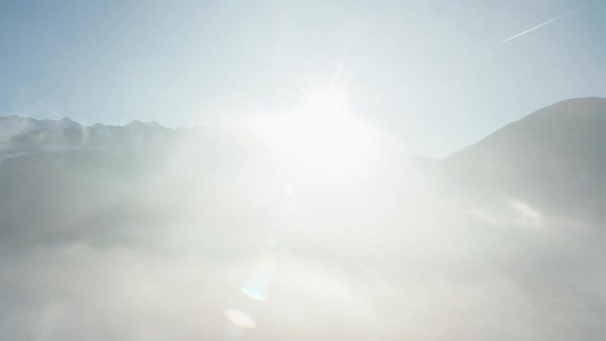 A helicopter flying next to clouds and mountains with the sun and the sky in the background