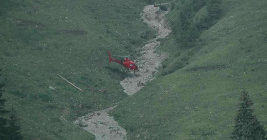 Red helicopter flies along dry stream bed away from person walking
