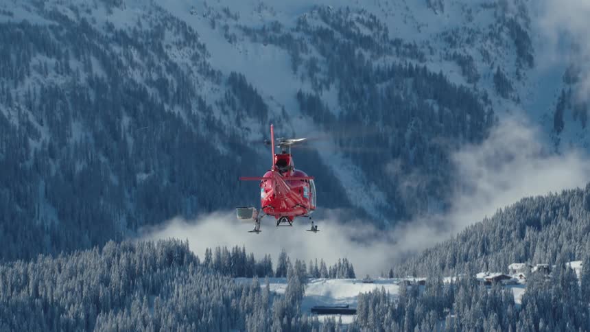 A helicopter following over a forest in the valley of a mountain range covered in snow