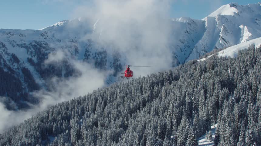Red helicopter flies over the trees on the snow covered mountain