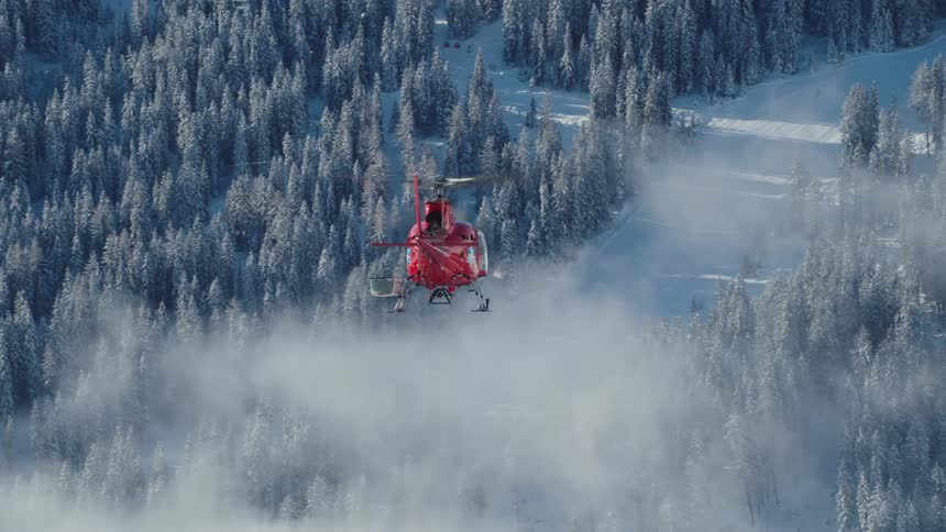 Red helicopter flying through a forest and above fog