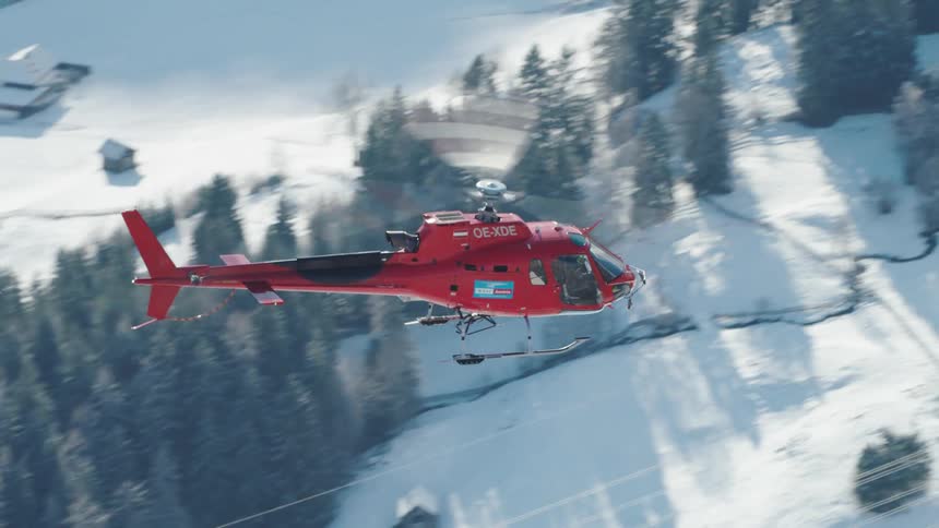 Red helicopter flies over snow covered mountain passing town