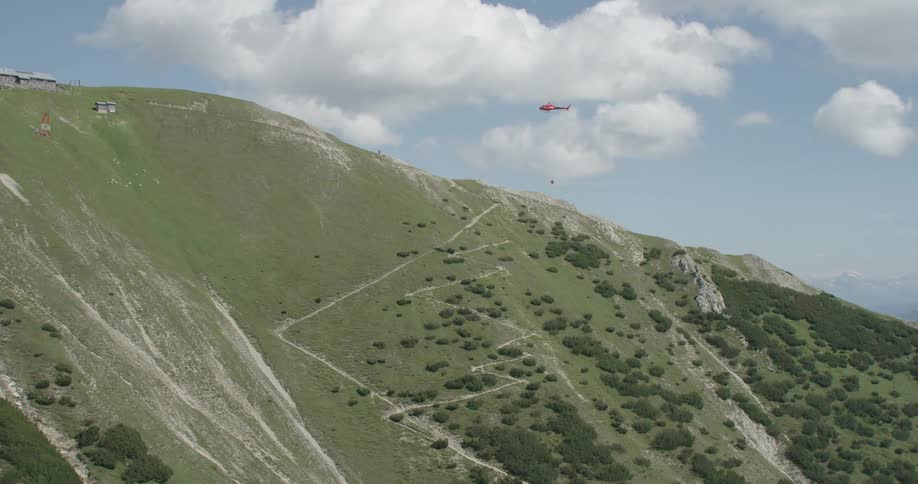 A helicopter carrying a water bucket flies over a green ridge and slope