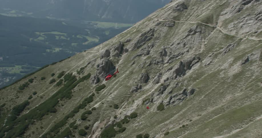 Red helicopter flies over the mountains while carrying a basket