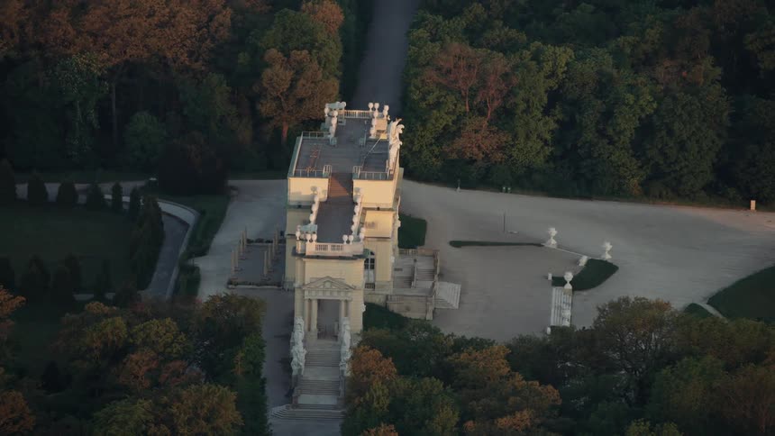 Sun touches top of ornate mansion surrounded by forest where tree foliage is just starting to change colors