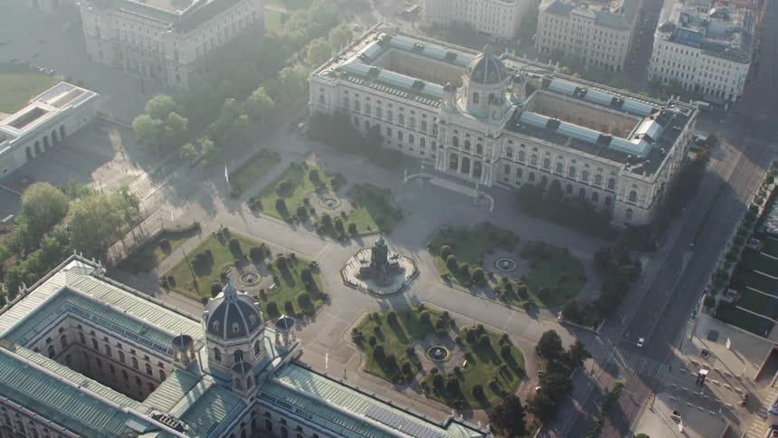 Cars travel on a city street past palaces and ornate buildings with an empty square