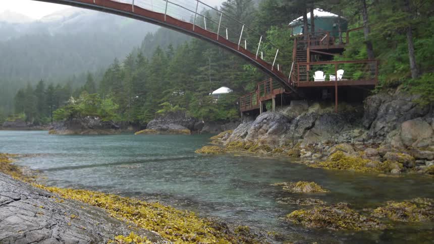 A river flows under an arched pedestrian bridge which leads to a wooden deck with adirondack chairs