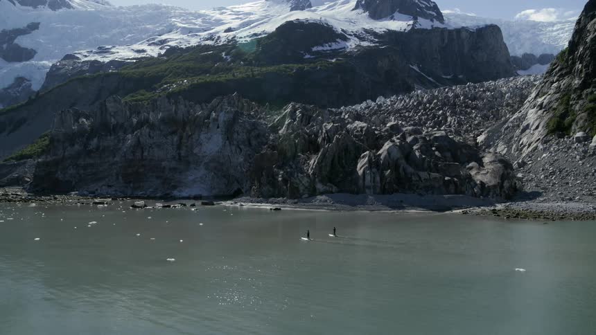 Two people stand on surfboards in the water among floating ice near mountains