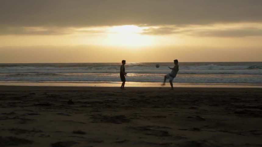 Two boys kick a ball on the beach at sunset