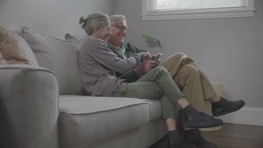 An elderly couple sit on the couch and laugh over viewing a cell phone