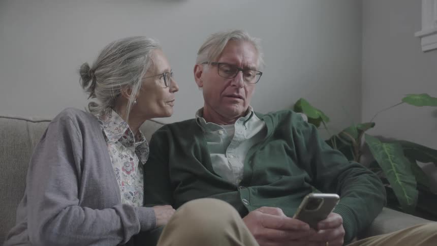 Elderly man and woman look at a cell phone as they sit on a sofa