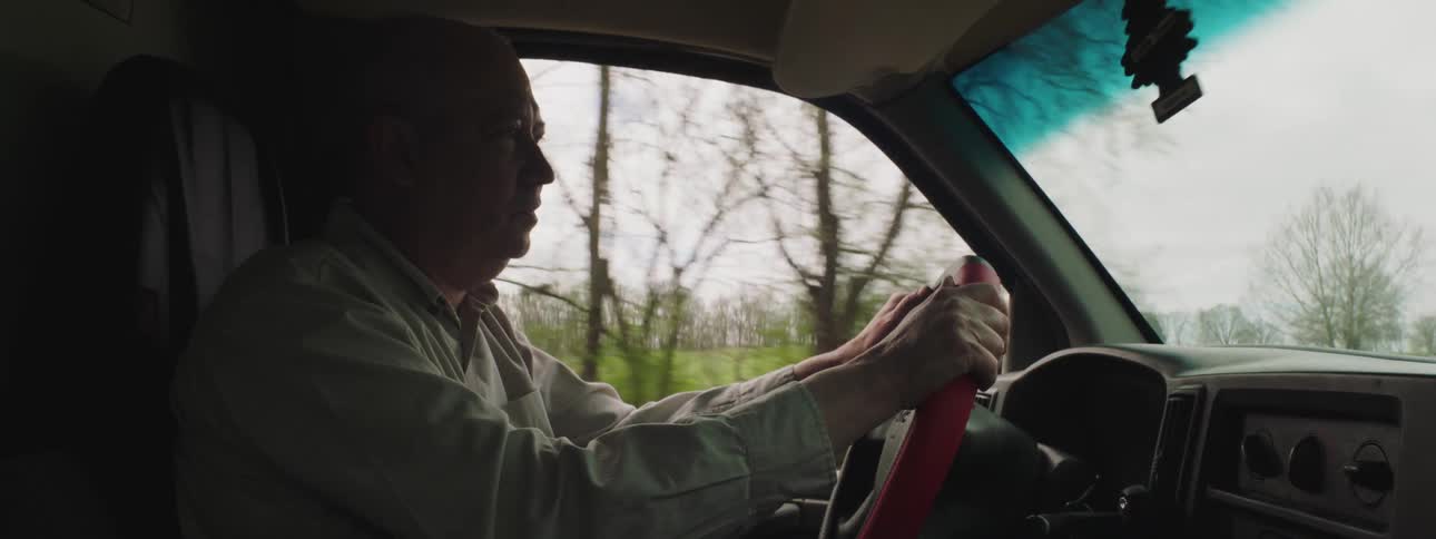 Male driving a truck with a red steering wheel along a road in a rural area