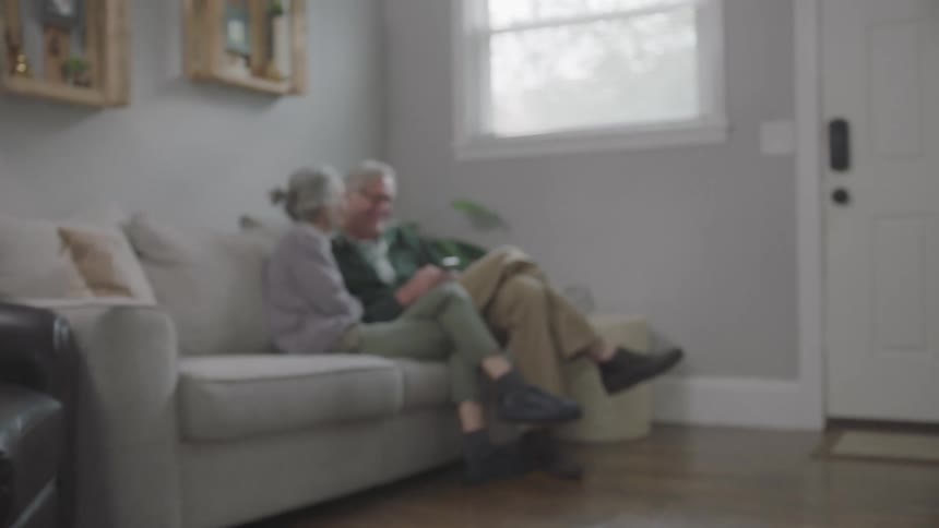 An elderly couple sit on a couch together looking at a cell phone