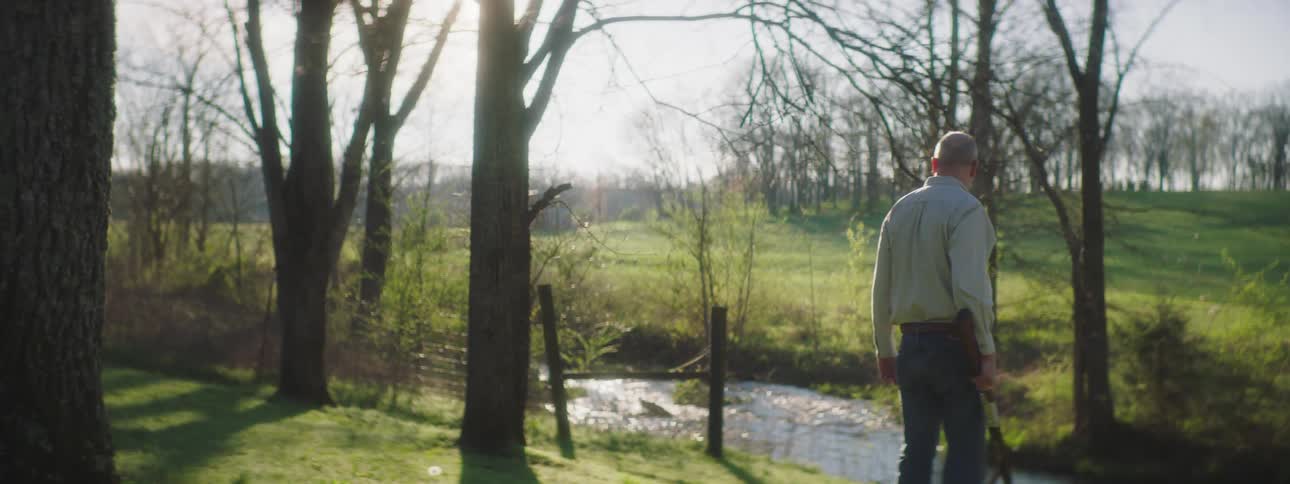 Man stands on shore near fence post looking down at stream