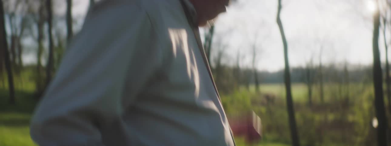 An older man walks through a forest with a rifle and coughs into his hand