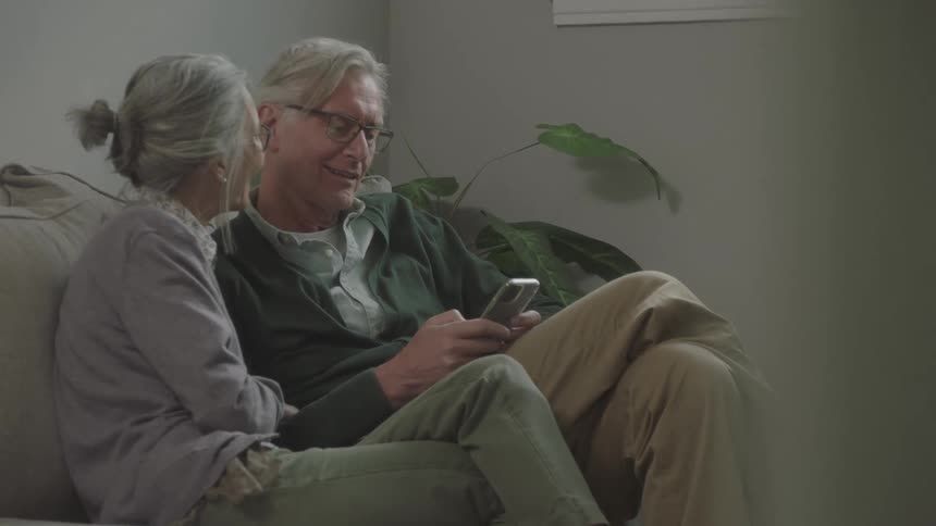 Elderly couple sitting on couch smiles and laughs using smartphone