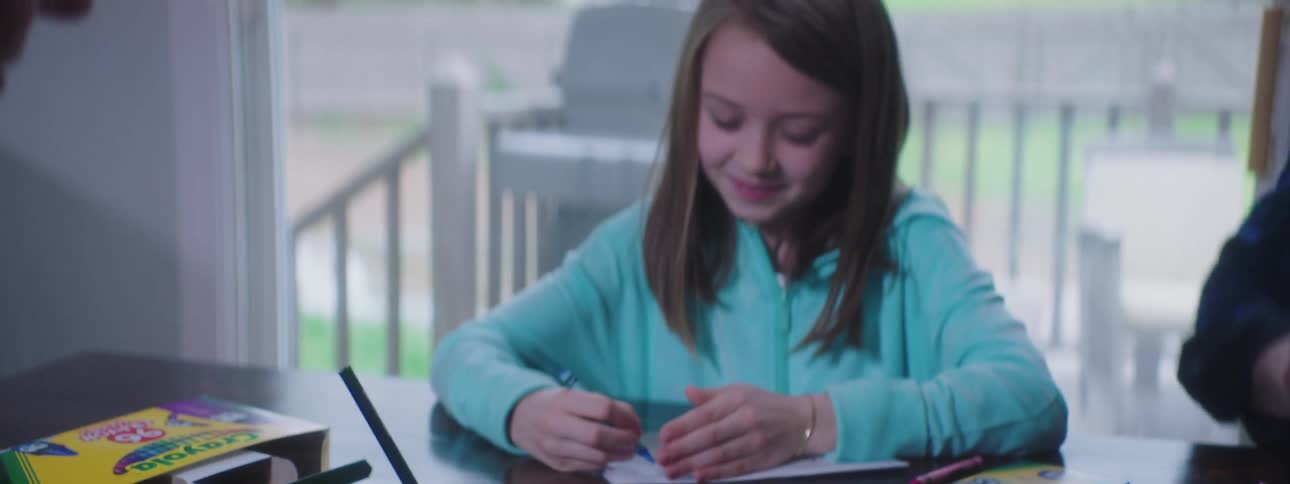 A girl using crayons to draw on a sheet of paper while sitting at a table with two other people who are also drawing