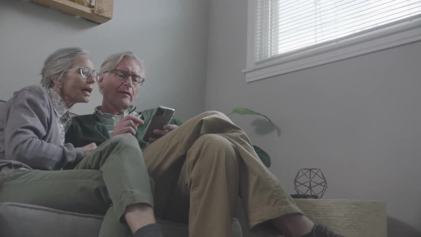 A mature couple are sitting on a sofa while interacting with a smartphone