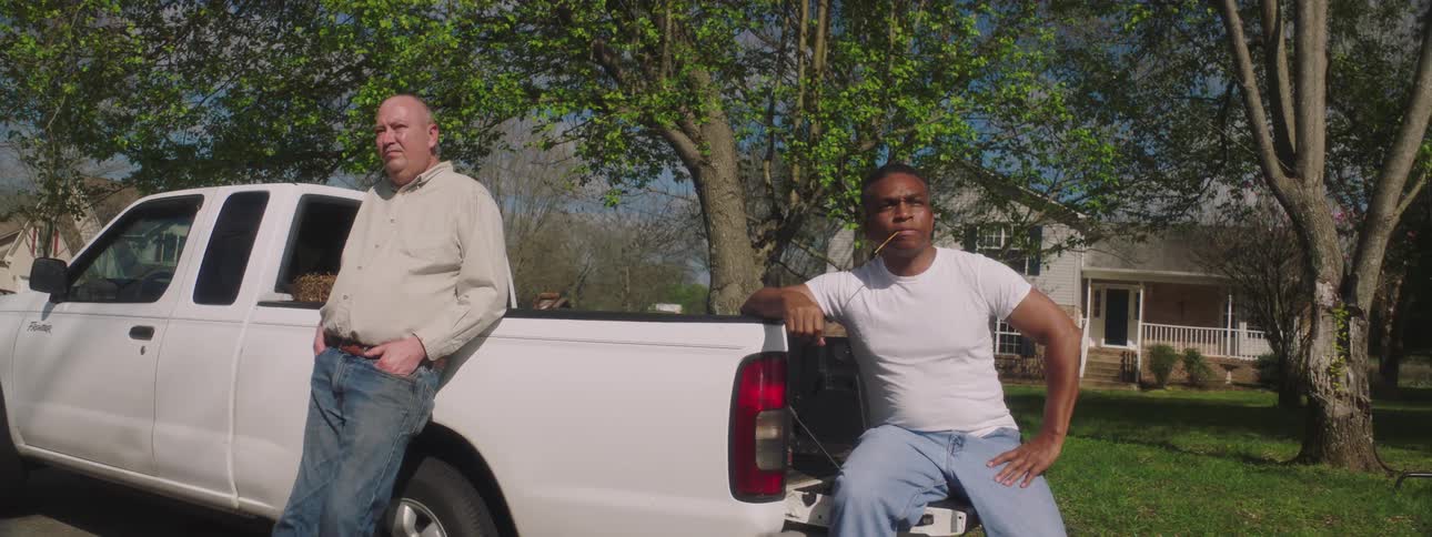 A white man leans against a truck and cough and a black man sits on the tailgate and looks over at him with concern