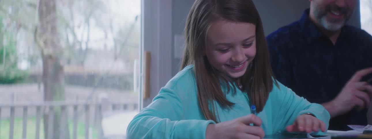 A girl drawing on a sheet of paper with a crayon while sitting at a table next to her dad
