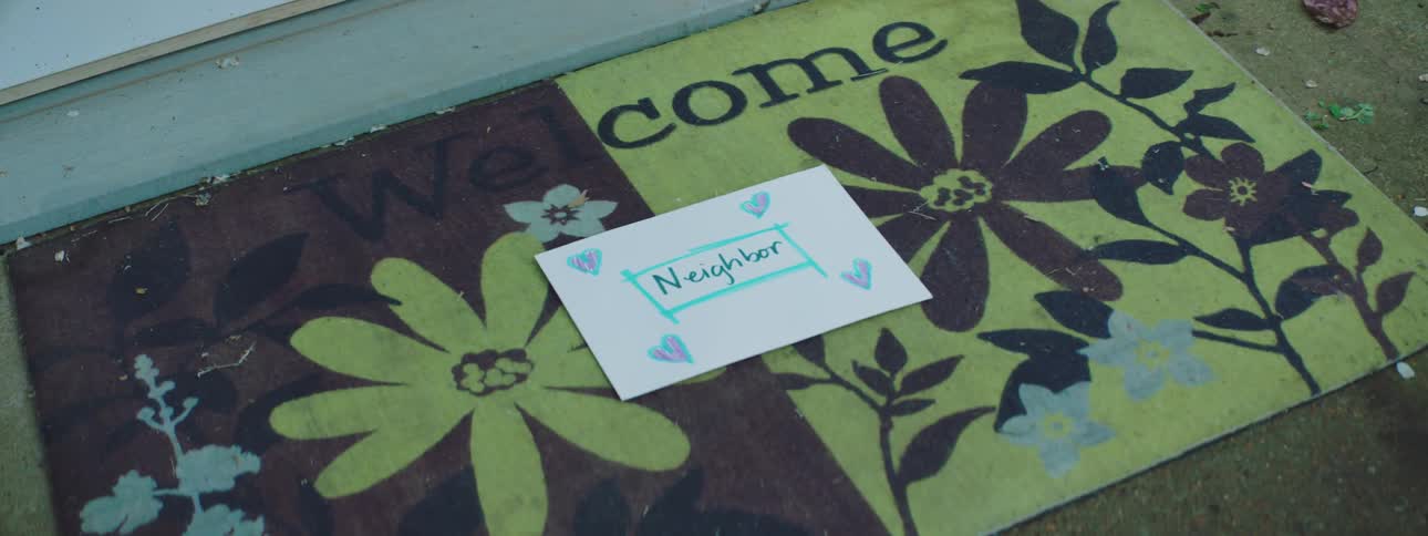 A card envelope addressed to neighbor and decorated with hearts is dropped on the welcome mat of a home