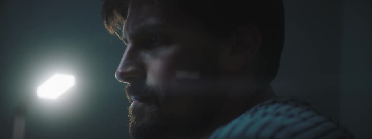 A man sits and licks his lips in a dimly lit room