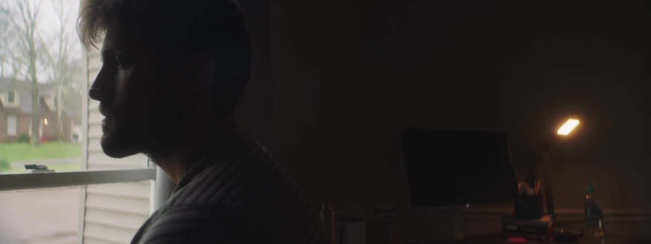 Bearded man in a dim lit room looks outside through window