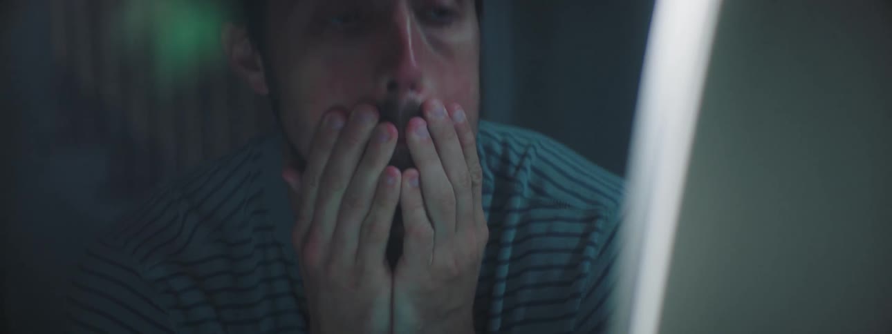 Man feeling tired and overwhelmed sits at the desk and uses the desktop in a lim lit room