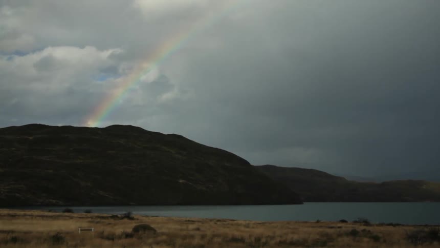 A rainbow can be seen in the distance behind a mountain by the beach