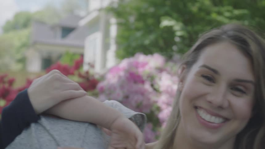 A mom sitting outside in her yard holds up her young son and he flops backwards as she laughs and shakes her head