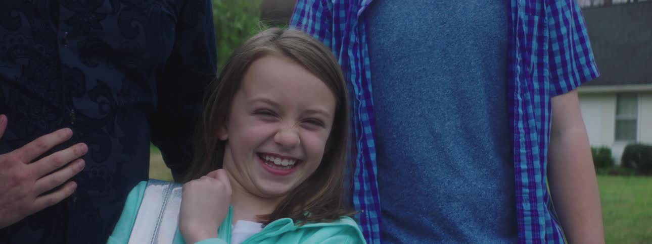 Girl with family smiles and laughs at the camera in the lawn of a house