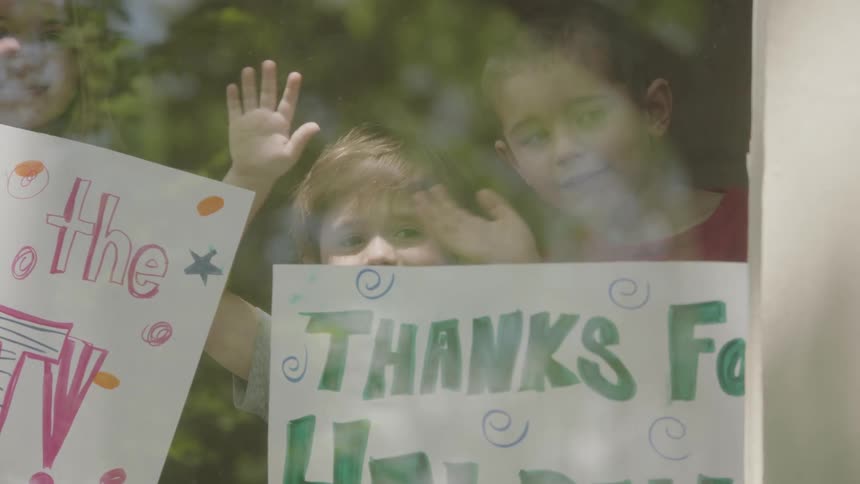 Three children hold signs with a thank you message as they wave out the window