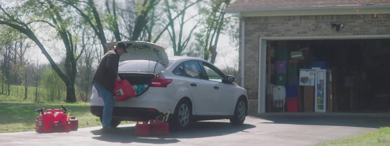 A man opens the trunk of his car and unloads gas and water cans before carrying packages of toilet paper into his garage