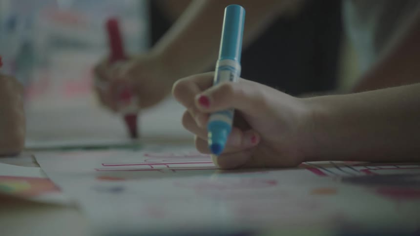 Children lean over table to draw on white paper with colored markers
