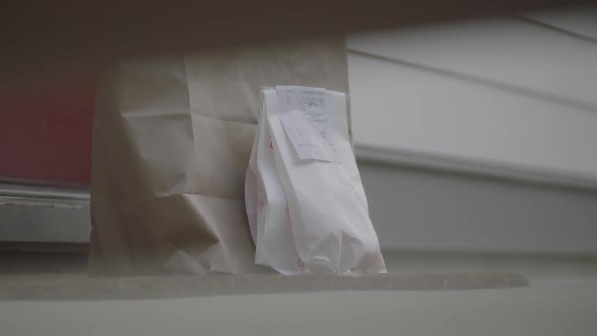 A woman drops off a bag of groceries and a prescription bag and walks away before the door opens and a person brings it inside