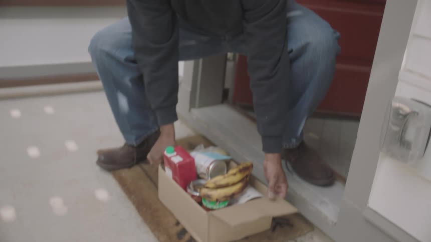A man opens his door waves and bends down to pick up the groceries that were delivered to him