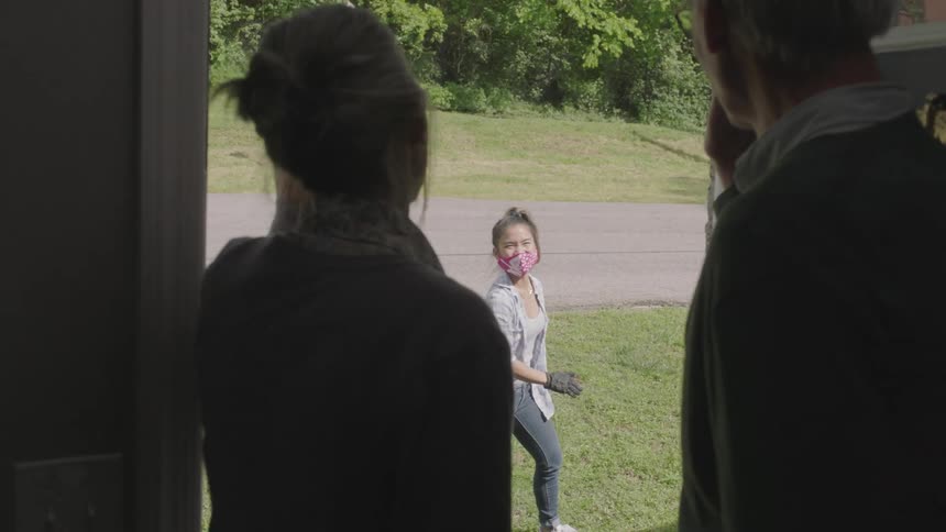 Older couple opening door to a young female wearing a mask waving from a distance