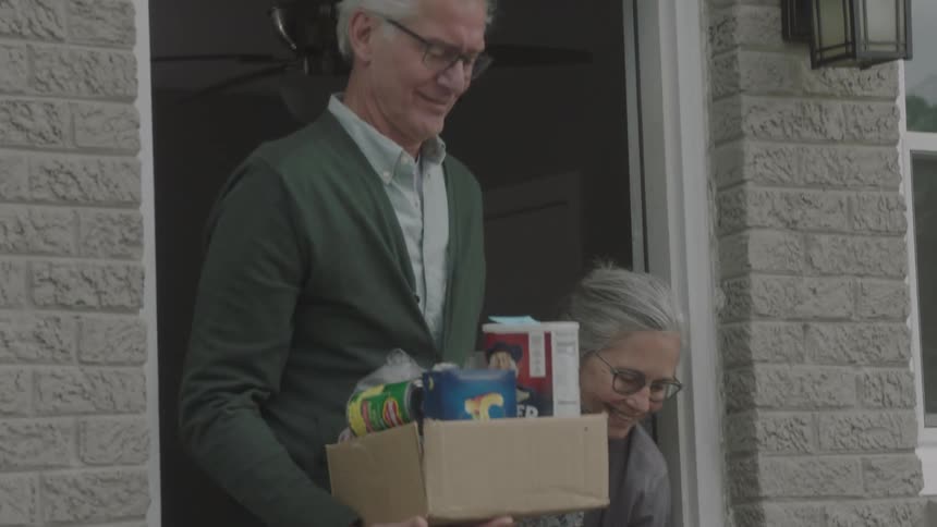 A man and woman stand in their doorway and pick up a box of groceries from their front step to take inside