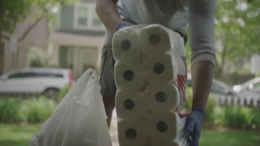 A delivery man wearing a mask and gloves carries groceries up to a front porch and leaves them on the step