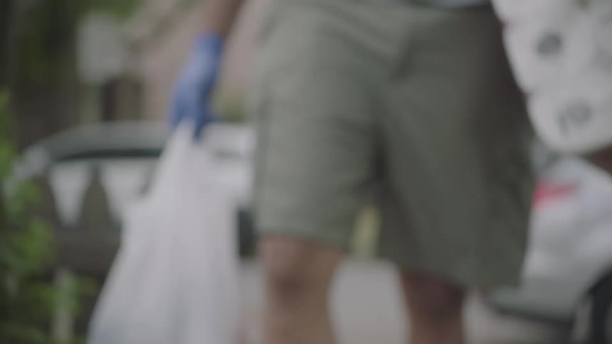A man walks down a sidewalk towards the camera holding groceries and wearing a blue glove