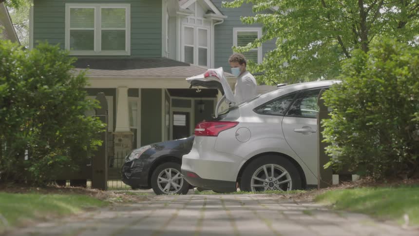 A man walking to the trunk of a car and retrieving a plastic bag and a large package of tissues