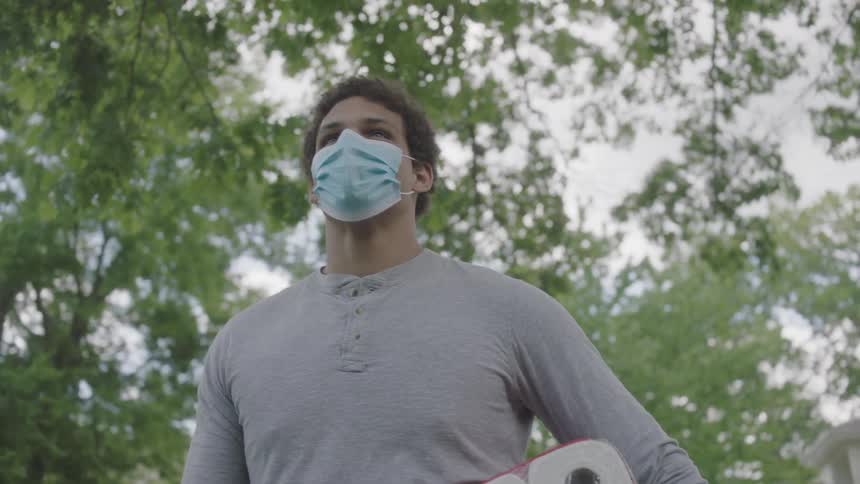 A man wearing a protective mask and gloves stands on a walkway holding a grcoery bag and a large package of toilet paper