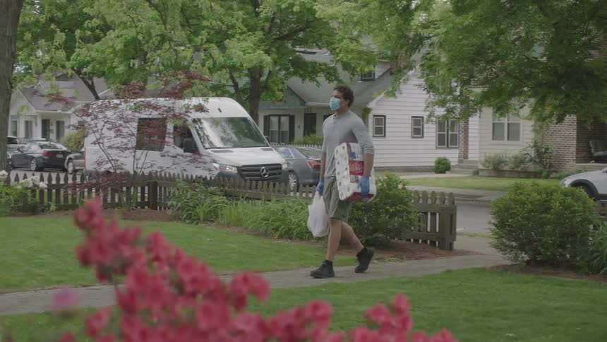 A delivery man wearing a mask and gloves carries groceries up a home sidewalk