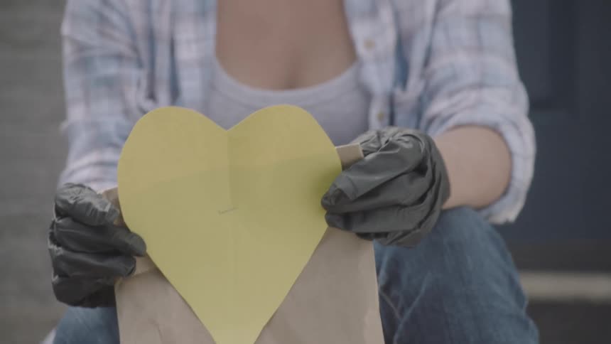 Female sitting on front porch wearing a face mask and gloves while holding a paper bag with a large yellow heart cut from construction paper