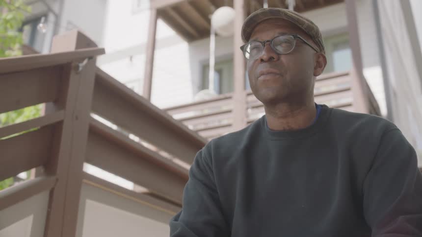 A black man sits outside and looks down at his hands in thought and then looks ahead and smiles