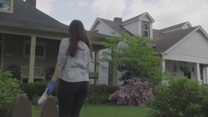 Woman wearing surgical mask and gloves delivers groceries to a front porch
