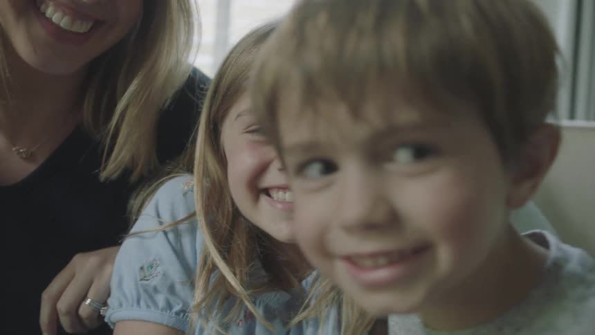 Two children smile for the camera while a third child pushes one of them and the mother watches and laughs